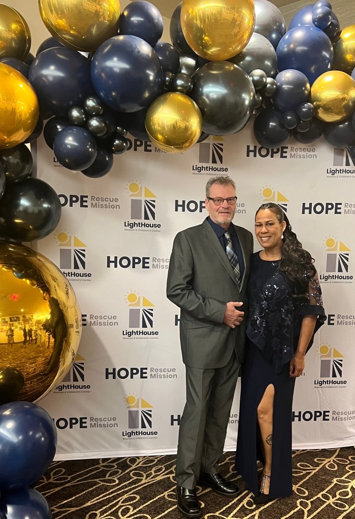 A man and a woman pose for a picture in front of a large banner that has logos for two organizations: Hope Rescue Mission and Lighthouse. There are gold, black and grey balloons hanging around the banner. The man and the woman are wearing formal clothes.